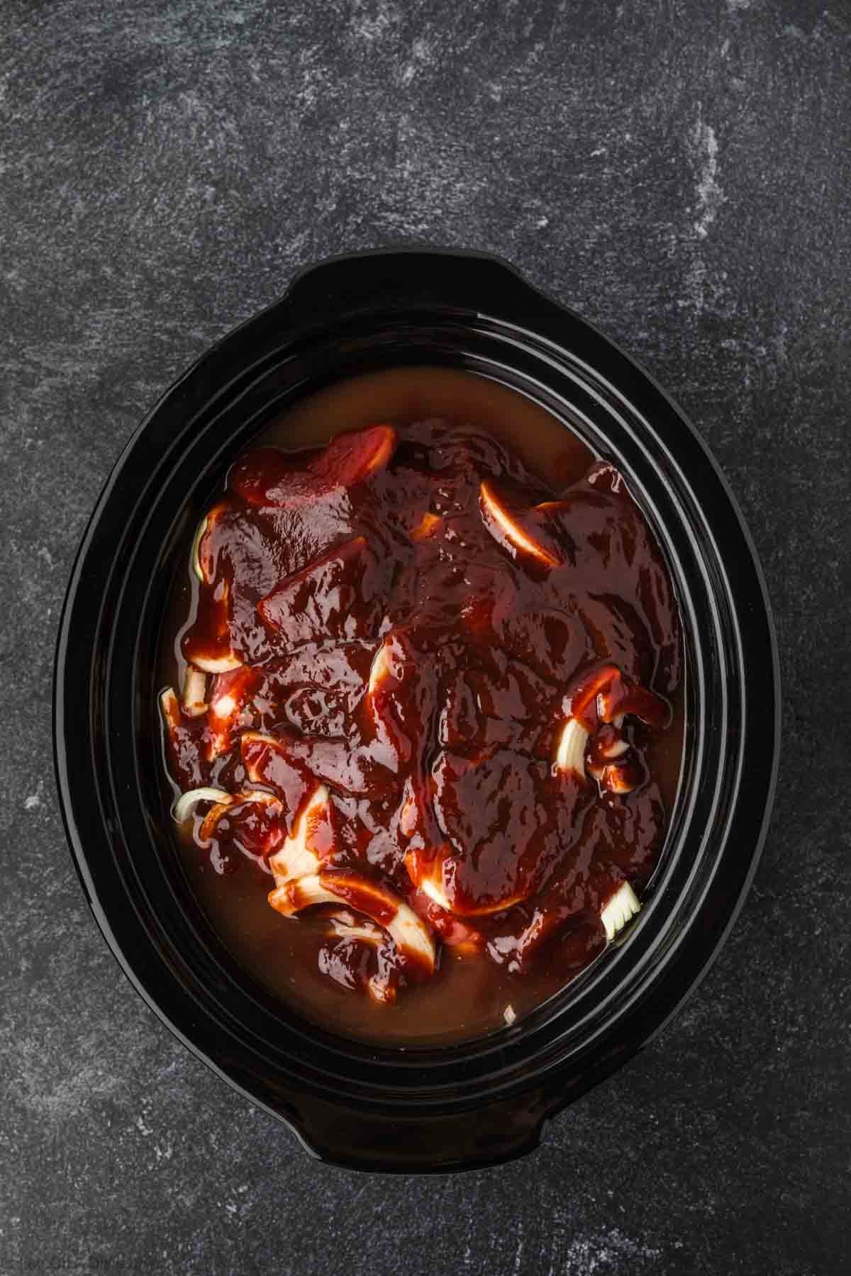 A slow cooker filled with raw meat, sliced onions, and barbecue sauce—perfect for a delicious pulled pork crock pot recipe—sits ready to be cooked on a dark textured surface.