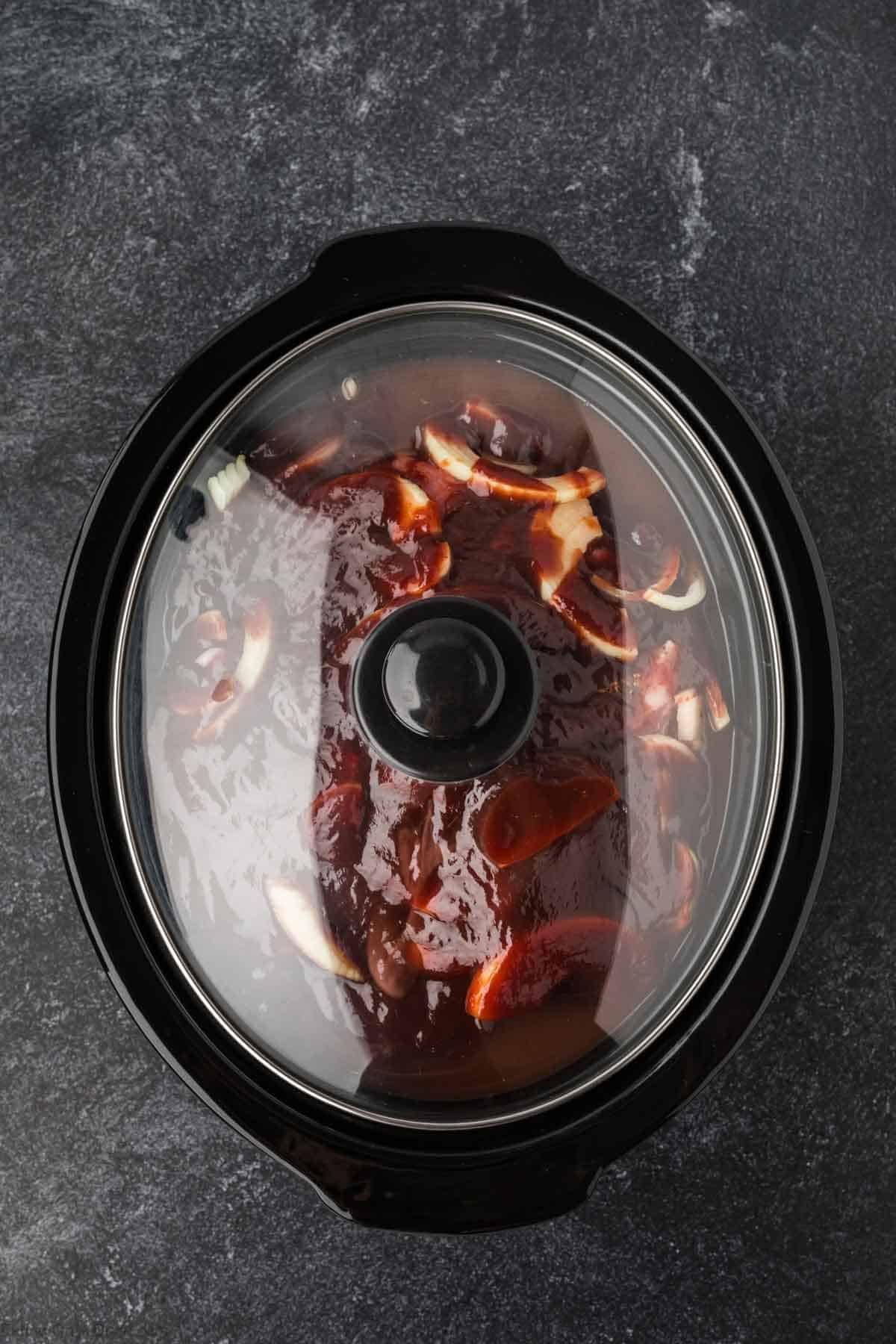 A slow cooker with a glass lid reveals a savory Pulled Pork Crock Pot Recipe, featuring a piece of meat smothered in barbecue sauce and sliced onions, set on a dark countertop.