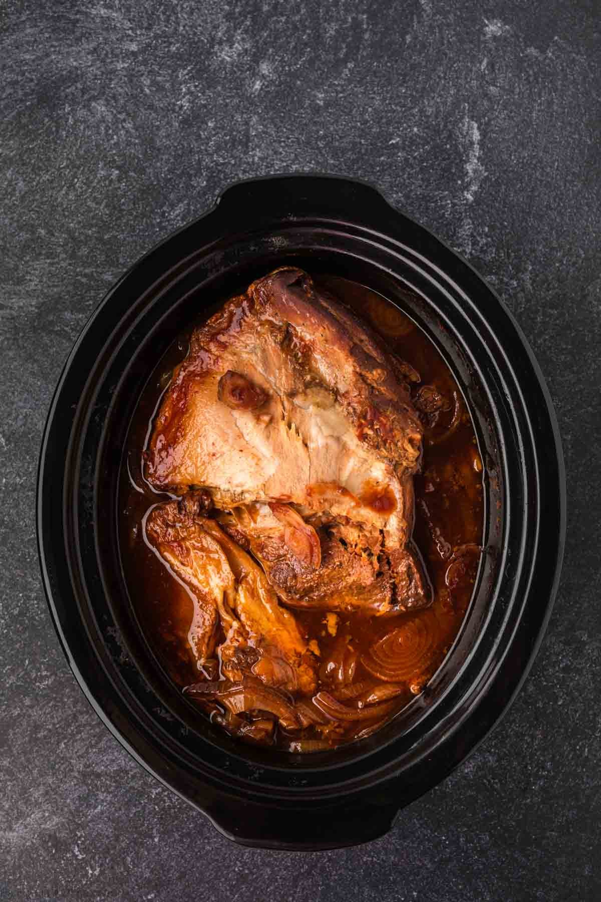 A cooked pork roast sits in a slow cooker surrounded by a rich, brown broth with visible onions, making it perfect for your favorite Pulled Pork Crock Pot Recipe on a dark textured surface.