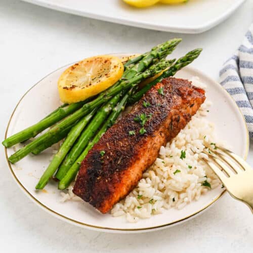 Blackened Salmon rests on a bed of white rice, accompanied by roasted asparagus and a grilled lemon slice, with a fork placed on the side of the plate.