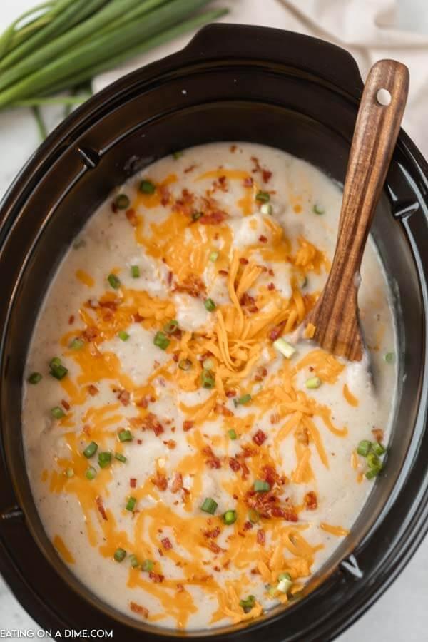 crock pot of potato soup with a wooden spoon and topped with shredded cheese, bacon and diced green onions