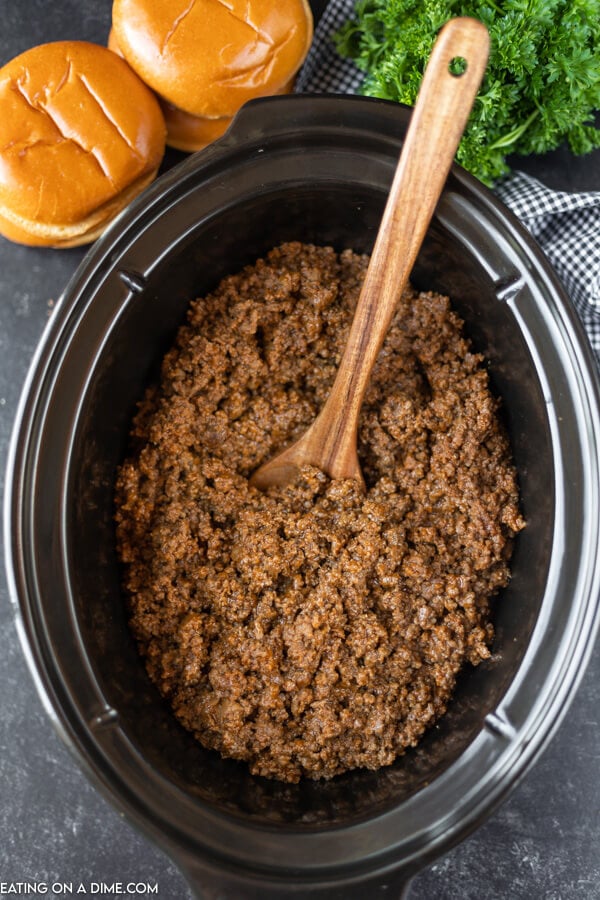 Close up image of sloppy joes in a crock pot with a brown spoons. Hamburger buns are also pictured next to the slow cooker. 