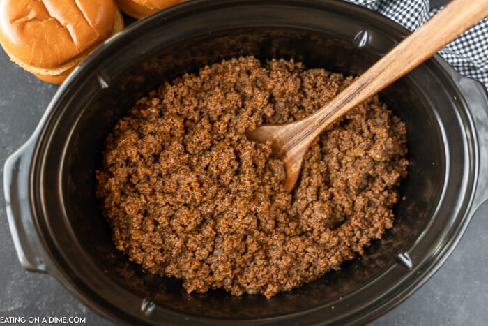 Close up image a sloppy joe mix in the crock pot with a brown spoon. Hamburger buns are on the side. 
