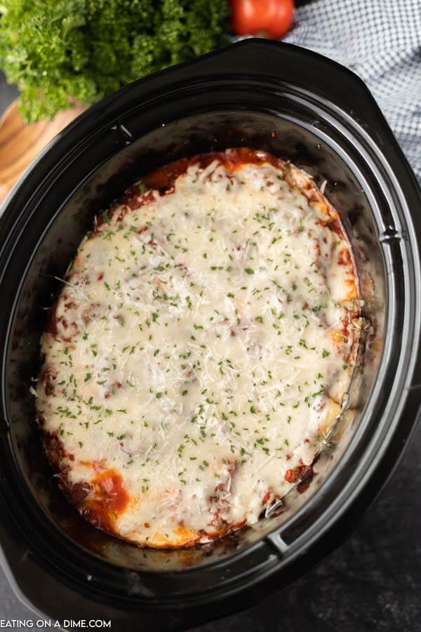Overview of this slow cooker lazy day lasagna recipe in a black crock pot with a large wooden spoon next to it and parsley behind it as well.  