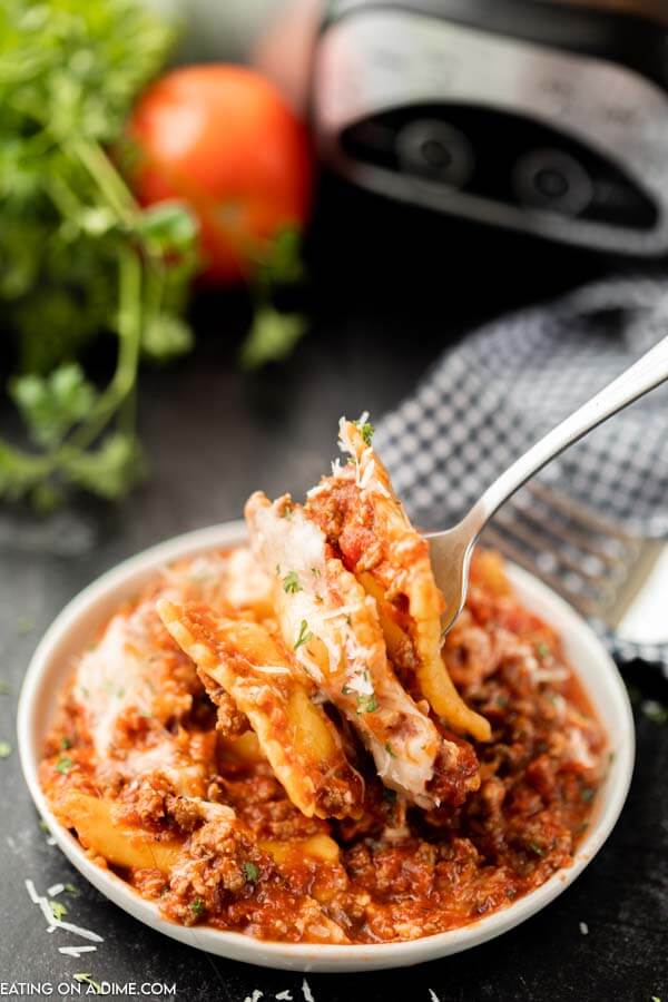 A fork with multiple raviolis on it lifting it away from the plate filled with a serving of this Lazy Day Lasagna with a crock pot, tomato and parsley behind it. 