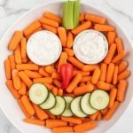 A playful Pumpkin Veggie Tray featuring baby carrots in a circle, two ranch dip cups as eyes, a red pepper nose, cucumber slice mouth, and celery stick eyebrows, all arranged to form a smiling face.