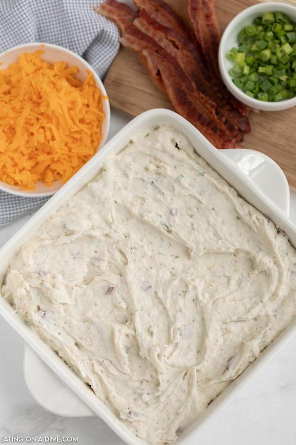 The mashed potatoes spread out in a casserole dish with bowls of cheese, bacon and chives next to it. 