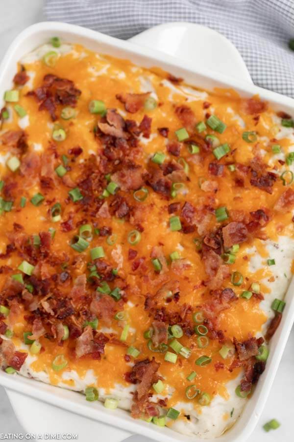 Close up of the loaded mashed potatoes casserole in a white casserole dish topped with cheese, bacon and chives with a plaid towel behind it. 