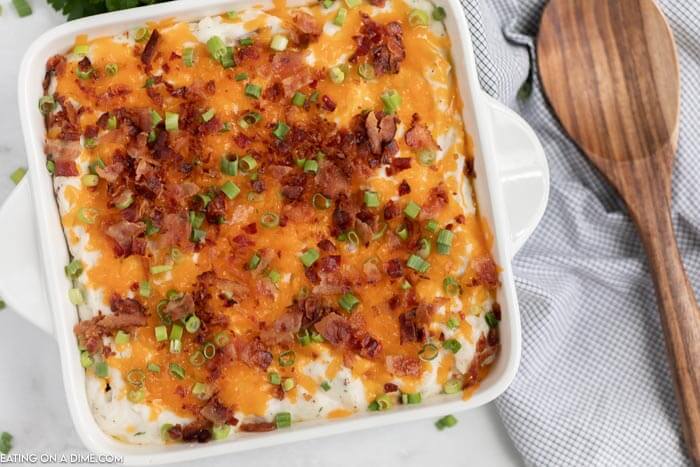 Close up of the loaded mashed potatoes casserole in a white casserole dish topped with cheese, bacon and chives with a wooden spoon next to it. 