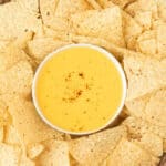 A white bowl of creamy yellow homemade nacho cheese sauce, sprinkled with a bit of spice, is surrounded by a layer of crispy tortilla chips.