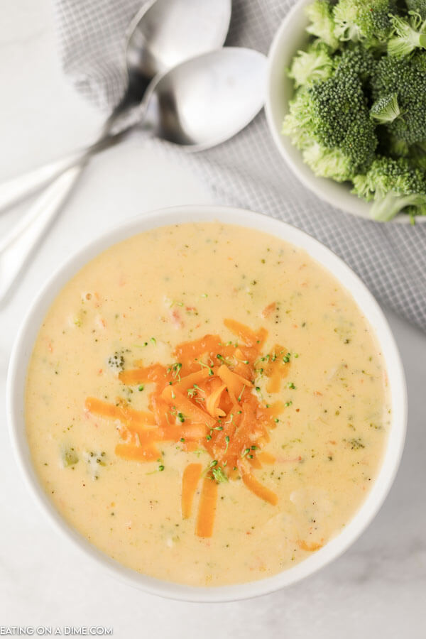White bowl of Panera Bread Broccoli Cheese Soup topped with shredded cheddar cheese with spoons and broccoli behind it. 