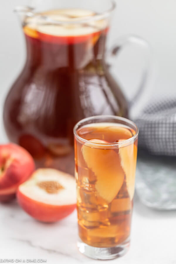 Close up image of a glass of tea with peaches in it. Also a pitcher of tea with peaches on the side. 