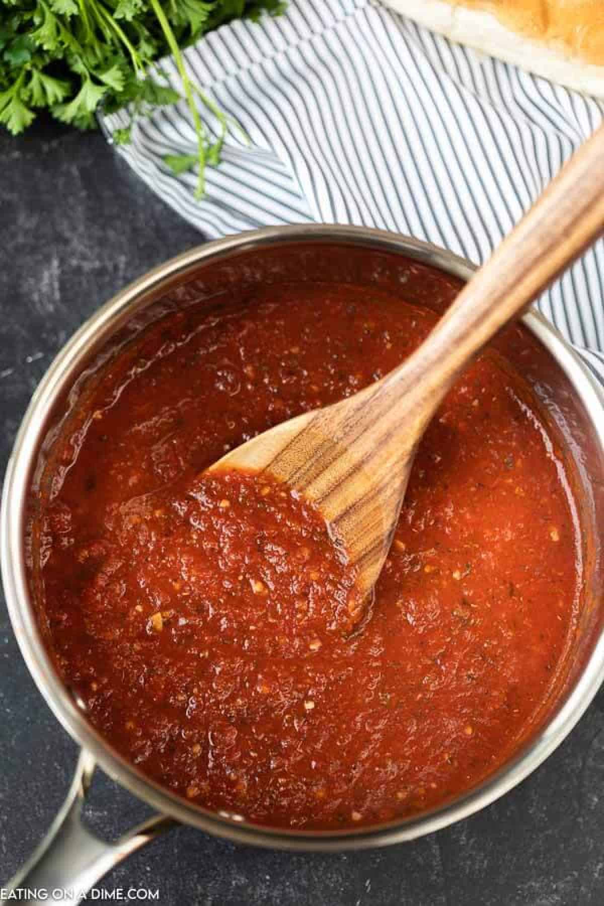A large wooden spoon scooping marinara sauce out of a metal saucepan. 