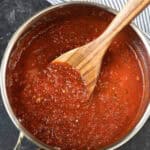 A wooden spoon stirs thick, red homemade marinara sauce recipe in a stainless steel pot on a dark surface.