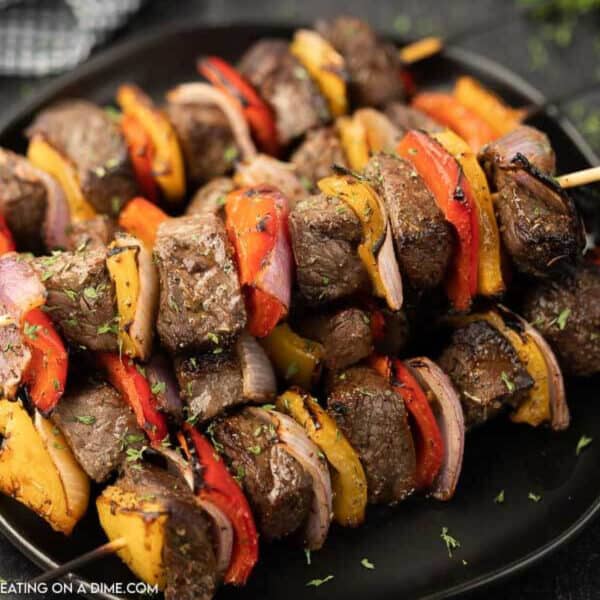 A close-up of shish kabobs on skewers with chunks of grilled beef, red and yellow bell peppers, and red onions, served on a black plate and garnished with herbs.