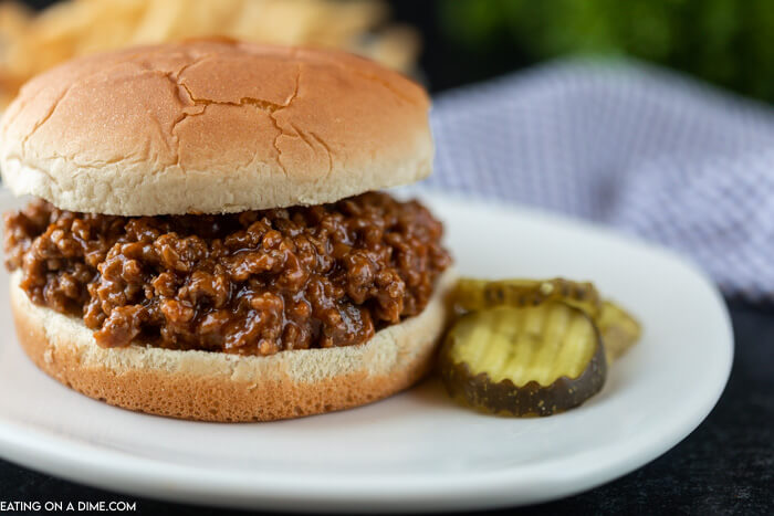 Close up image of sloppy joes on a hamburger bun with a side of pickles. 