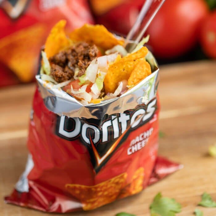 A close up of a walking taco with Doritos on a wooden platter with a plastic fork in the bag.