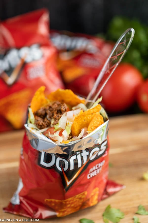 What is a walking taco? You are going to love this walking tacos recipe. It is easy to make and the entire family loves Walking tacos. Learn how to make a walking taco. We love making a walking taco bar for parties! A walking taco on a wooden platter with a plastic fork in the bag.