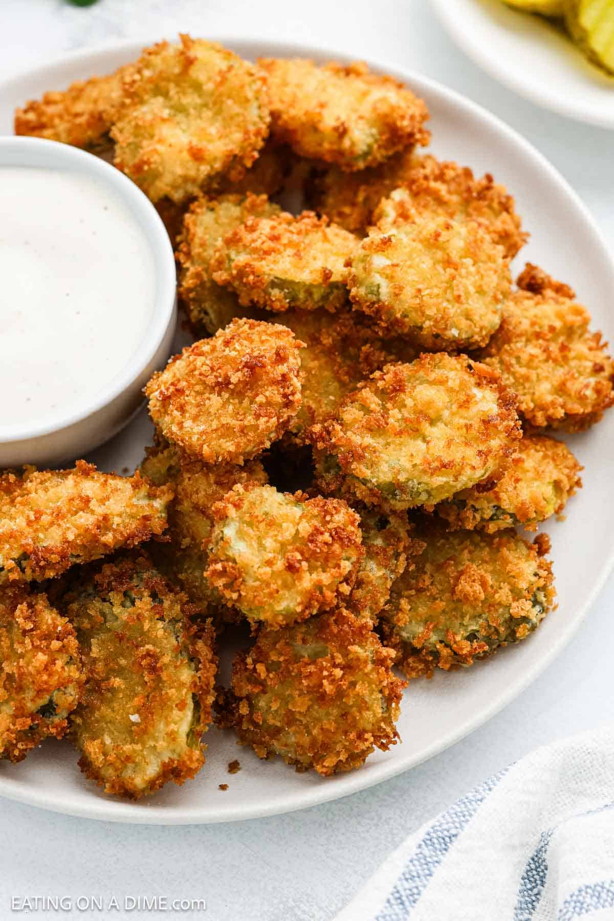 A plate of deep fried pickles coated in golden breadcrumbs is served with a cup of creamy dipping sauce on the side.
