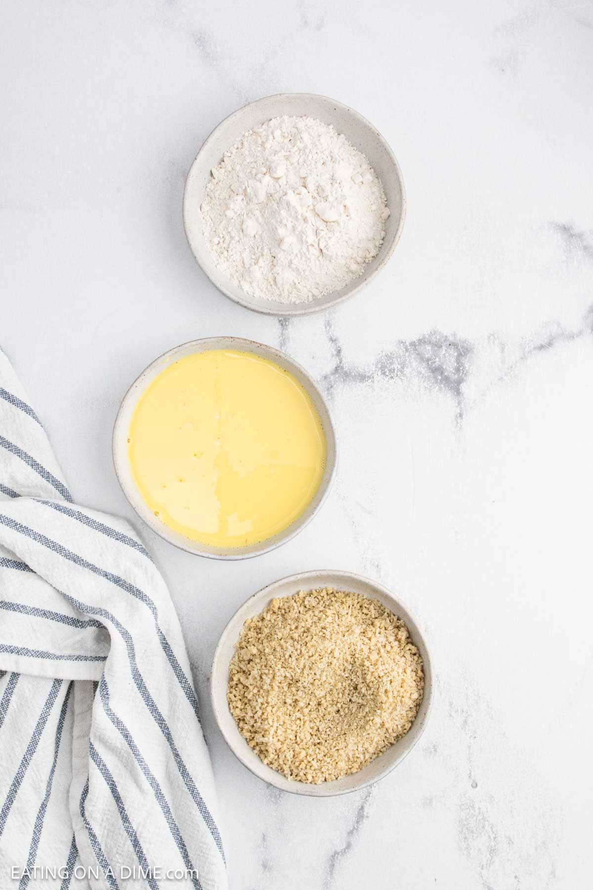Three small bowls with flour, beaten eggs, and breadcrumbs arranged vertically on a white marble surface next to a white and blue striped kitchen towel—perfect for prepping deep fried pickles.