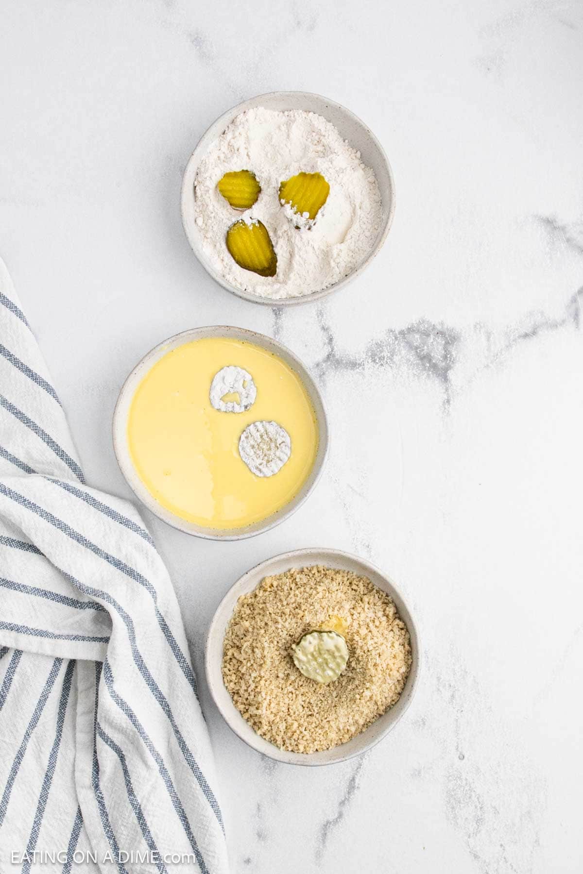 Three bowls show steps to breading deep fried pickles: flour with pickle slices, egg wash with flour-coated pickles, and breadcrumbs with an egg-coated pickle. A striped kitchen towel lies next to the bowls on a white countertop.