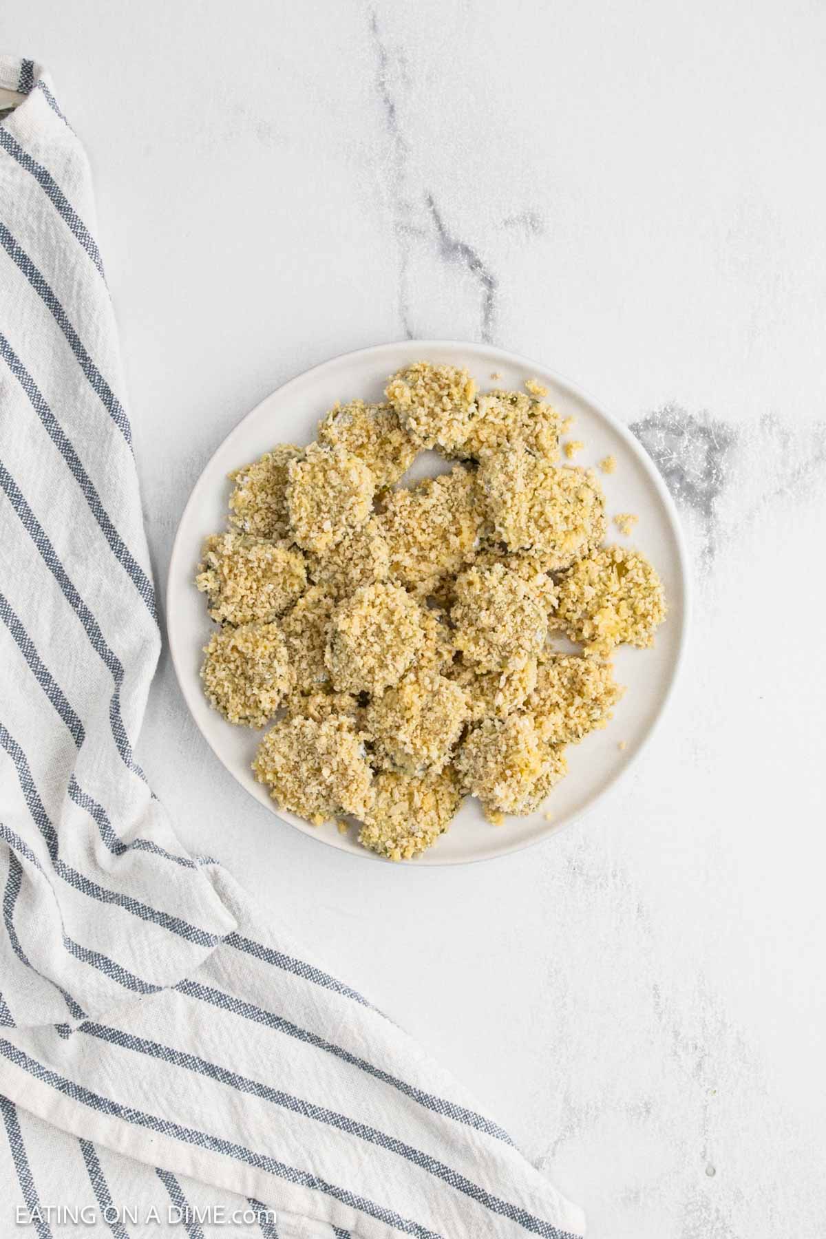 A white plate filled with breaded, uncooked deep fried pickles sits on a light surface next to a white and blue striped cloth.