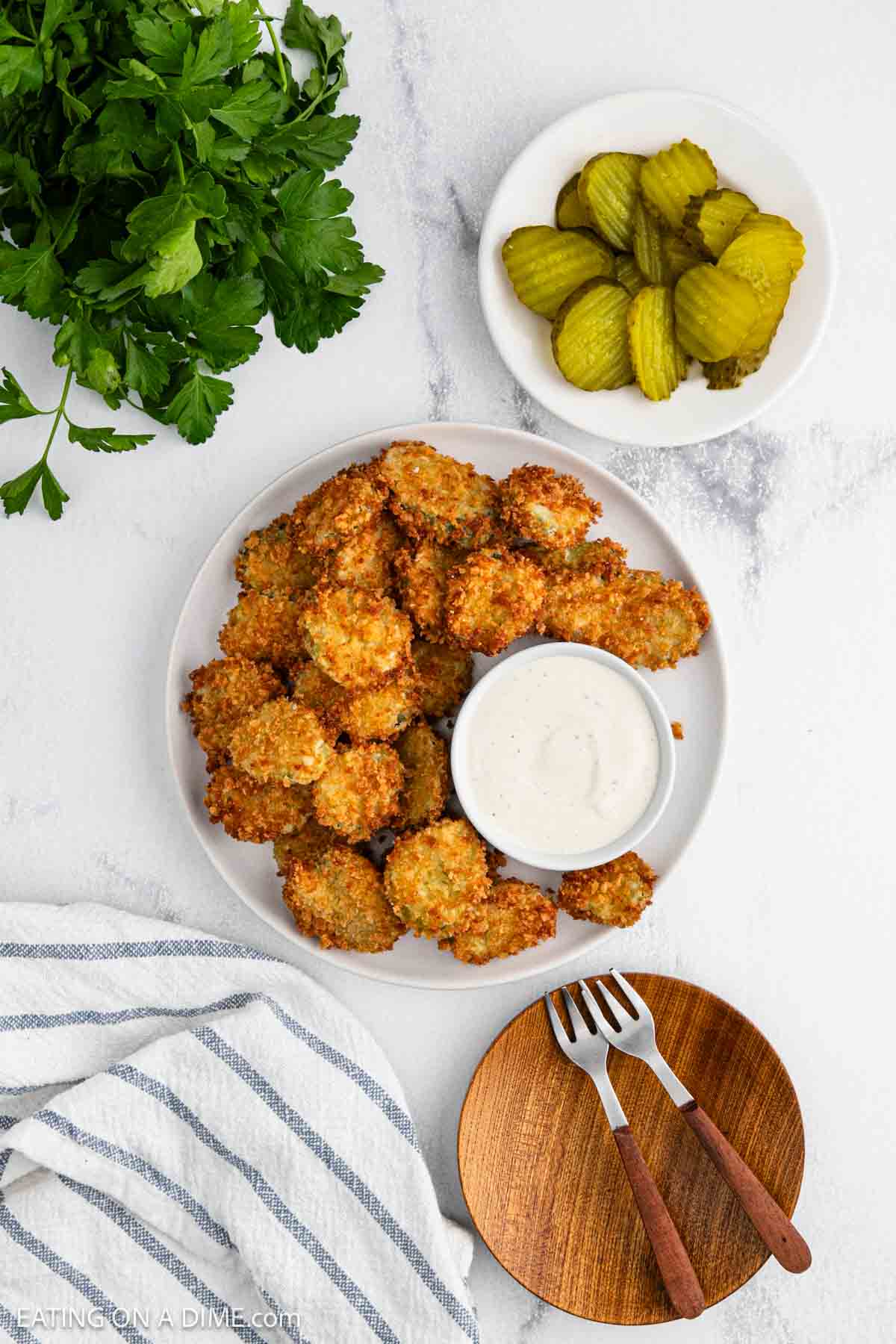 A plate of crispy, deep fried pickles is served with a bowl of ranch dipping sauce. Next to it are sliced pickles in a small dish, fresh parsley, and a striped napkin with two wooden plates and forks.