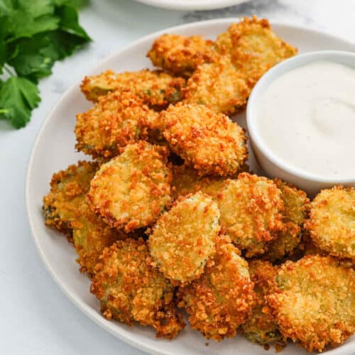 A plate of crispy, golden-brown deep fried pickles is served with a cup of creamy dipping sauce, with fresh parsley visible in the background.