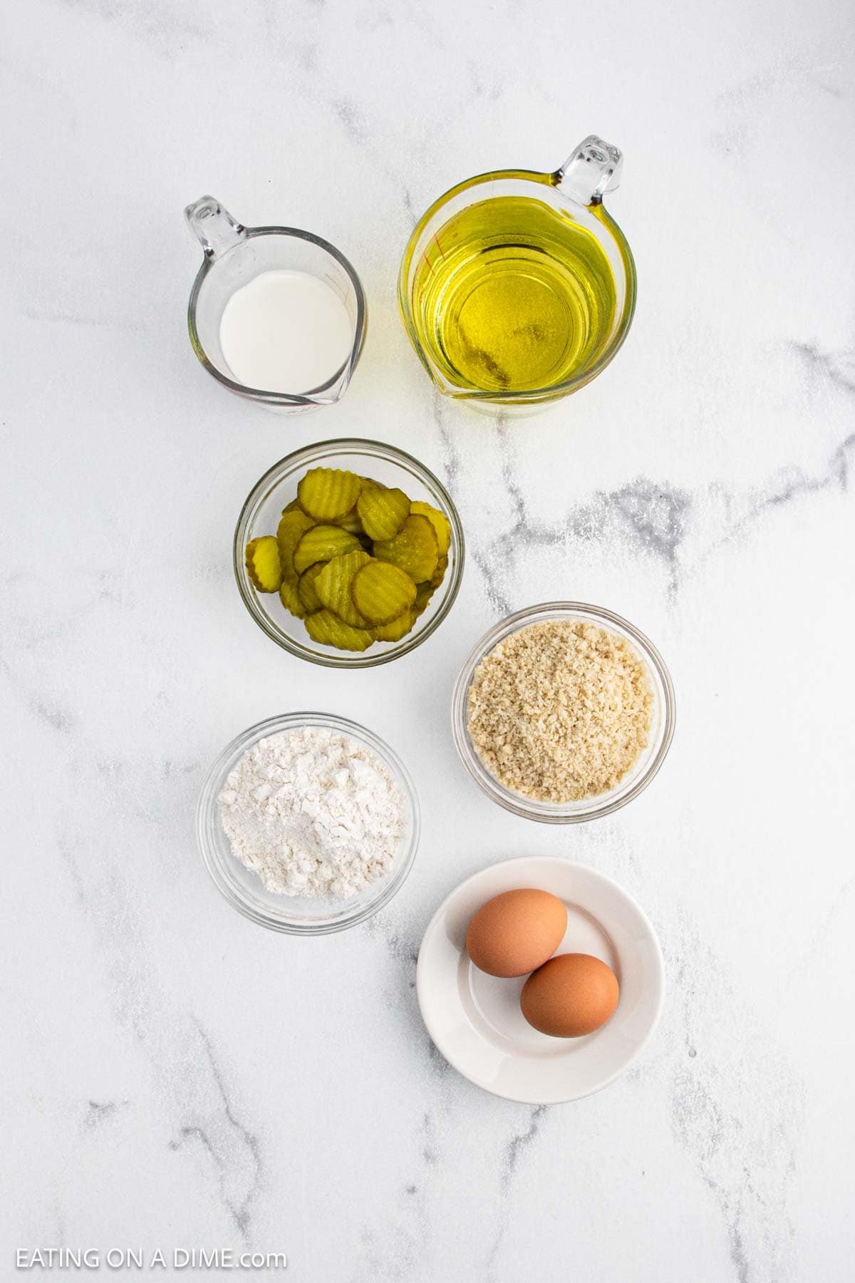 Six ingredients for deep fried pickles are arranged on a white surface: a cup of oil, a small cup of milk, a bowl of pickle slices, a bowl of breadcrumbs, a bowl of flour, and two brown eggs on a small white plate.