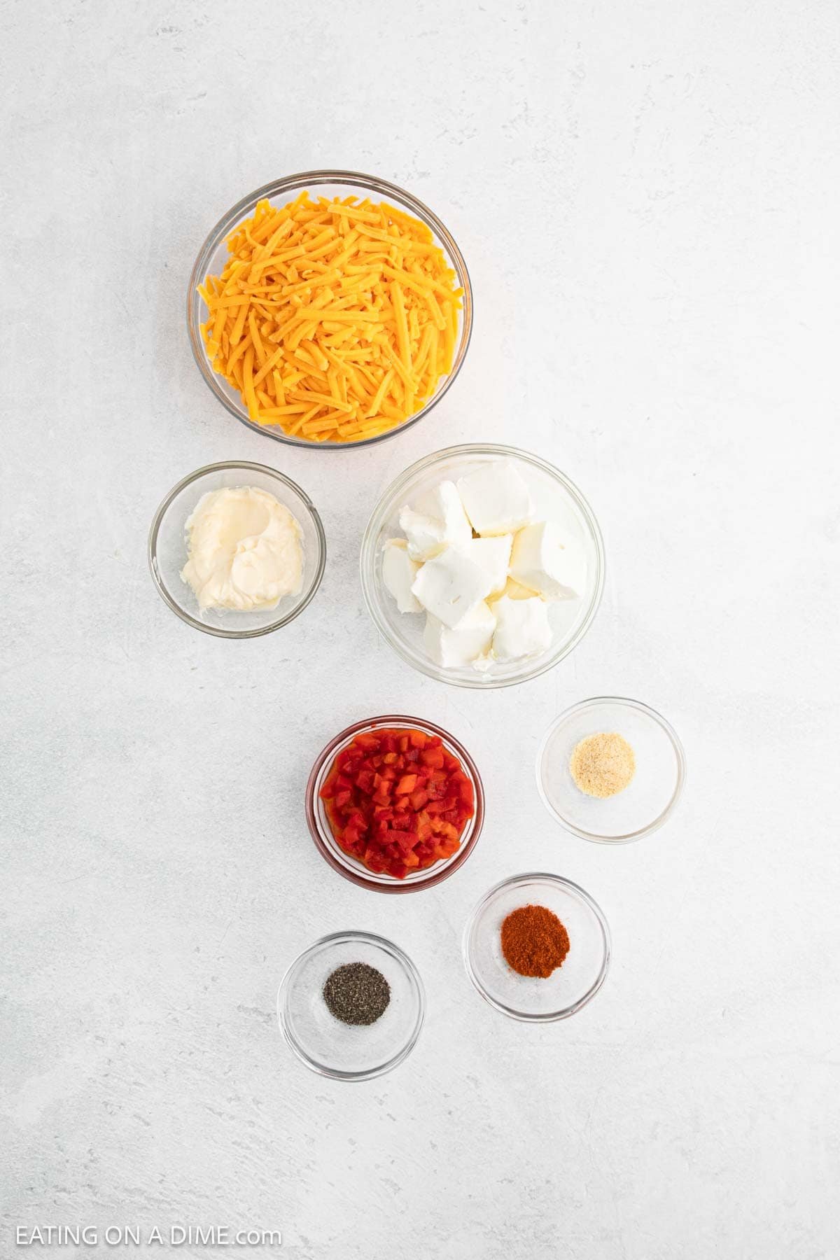 Six small glass bowls on a white surface hold ingredients for classic Pimento Cheese Dip: shredded cheddar cheese, mayonnaise, cream cheese, diced pimentos, ground black pepper, garlic powder, and paprika.