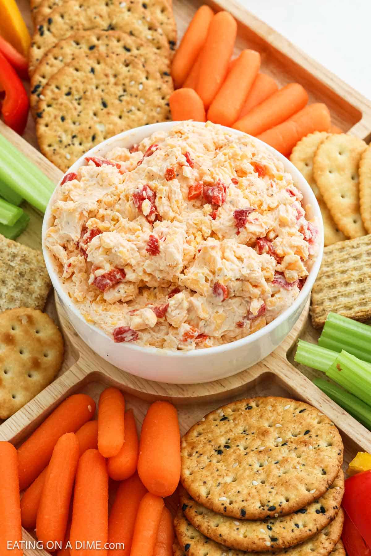 A bowl of creamy Pimento Cheese Dip is surrounded by assorted crackers, baby carrots, celery sticks, and red bell pepper slices on a wooden serving tray.