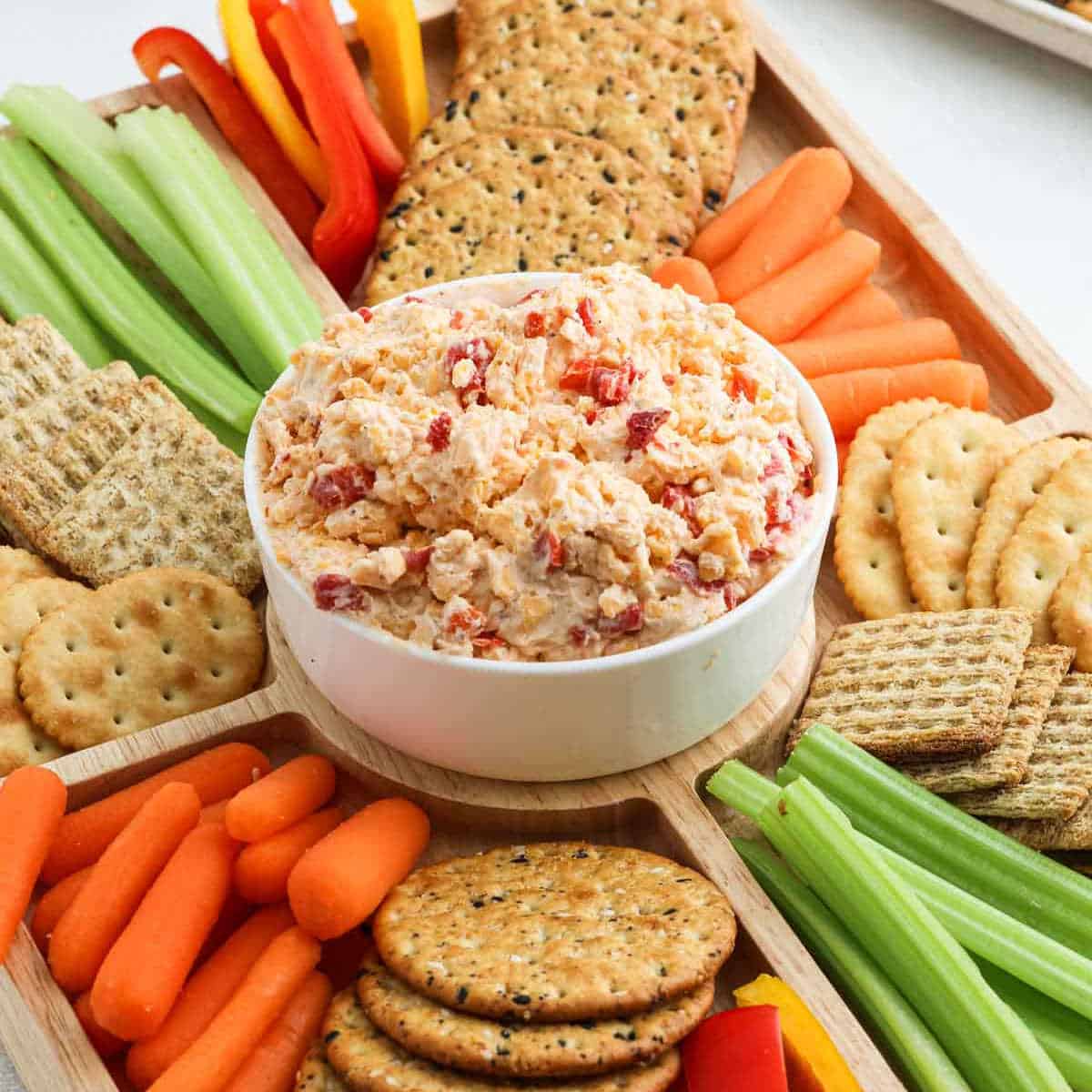 A wooden tray filled with assorted crackers, celery sticks, carrot sticks, and sliced bell peppers, centered around a bowl of chunky pimento cheese dip with red pepper pieces.