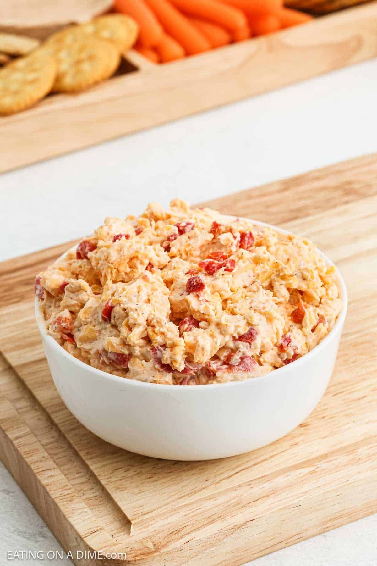 A white bowl filled with creamy Pimento Cheese Dip sits on a wooden cutting board. In the background, there are crackers and baby carrots on a wooden tray, perfect for pairing with this flavorful Southern classic.
