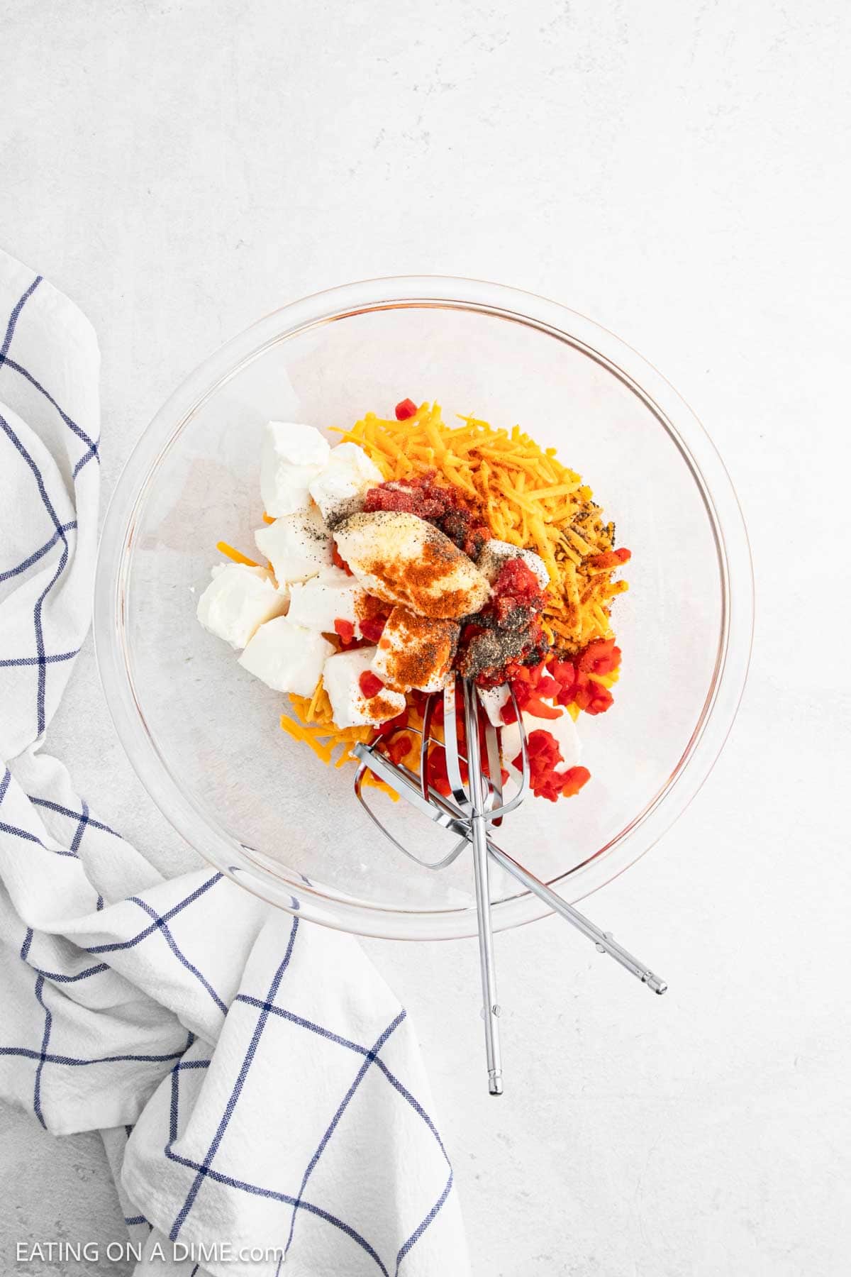 A glass bowl with shredded cheese, cream cheese, diced red peppers, and seasonings—perfect ingredients for a classic Pimento Cheese Dip. Two forks rest inside, next to a white kitchen towel with blue stripes on a light surface.