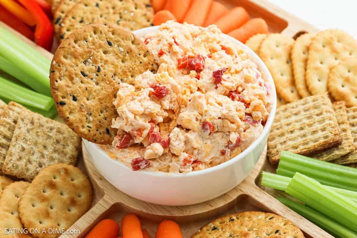 A bowl of creamy Pimento Cheese Dip sits on a wooden tray, surrounded by assorted crackers, celery sticks, and baby carrots. One cracker is dipped into the cheesy goodness.