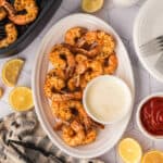 A white oval plate with seasoned Air Fryer Shrimp and a bowl of creamy dipping sauce, surrounded by lemon slices, ketchup, and utensils on a patterned table.