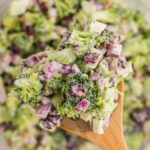 A close-up of a wooden spoon holding a creamy Broccoli Cranberry Salad with chopped red onions, raisins, and sunflower seeds, with more salad in the blurred background.