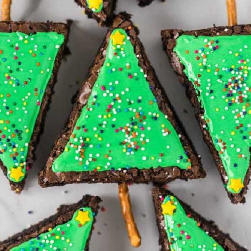 Christmas Tree Brownies slices decorated with green icing and colorful sprinkles to resemble festive trees, each with a pretzel stick trunk, arranged on a white surface.