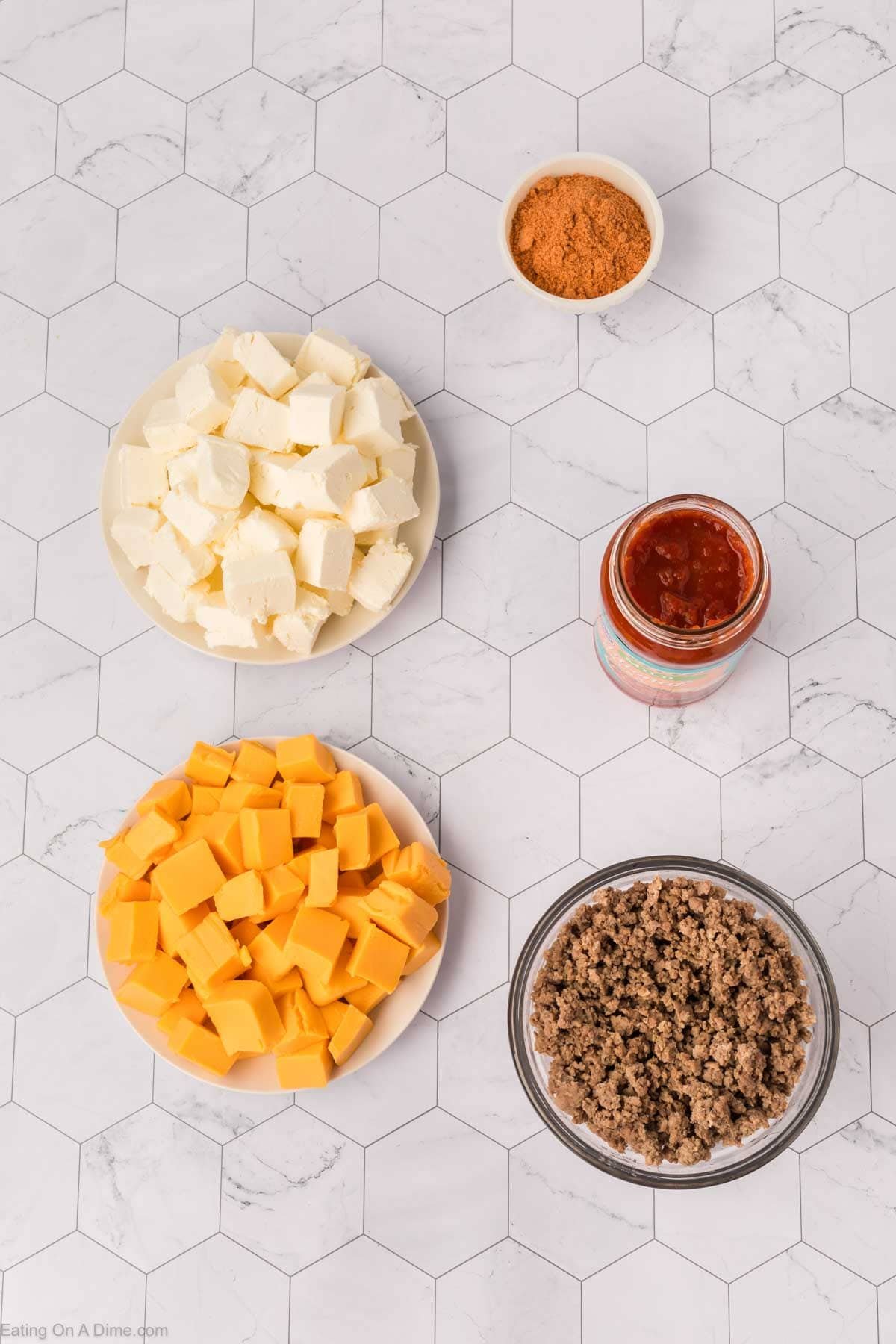 Overhead view of five ingredients for Crockpot Taco Dip on a white hexagon-tile surface: cream cheese cubes, taco seasoning, salsa, cooked ground beef, and cubed cheddar cheese.