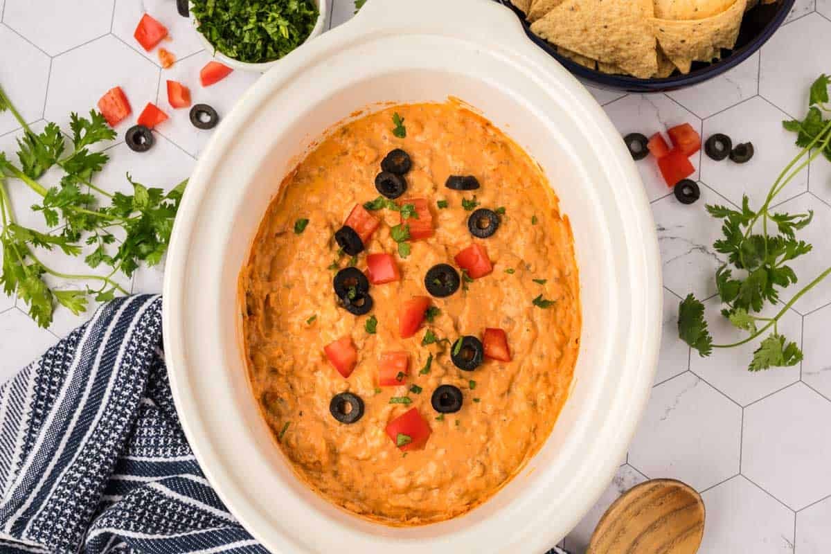 A white slow cooker of Crockpot Taco Dip, topped with chopped tomatoes, black olives, and parsley. Chopped herbs, tortilla chips, a striped towel, and scattered cilantro surround the dish on a white hexagonal surface.