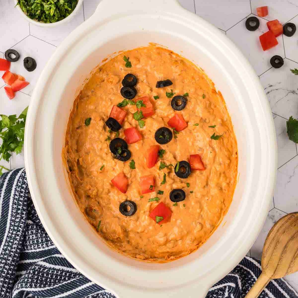 A creamy Crockpot Taco Dip in a white slow cooker, topped with sliced black olives, diced tomatoes, and chopped herbs. A blue and white striped towel and scattered ingredients surround the dish.