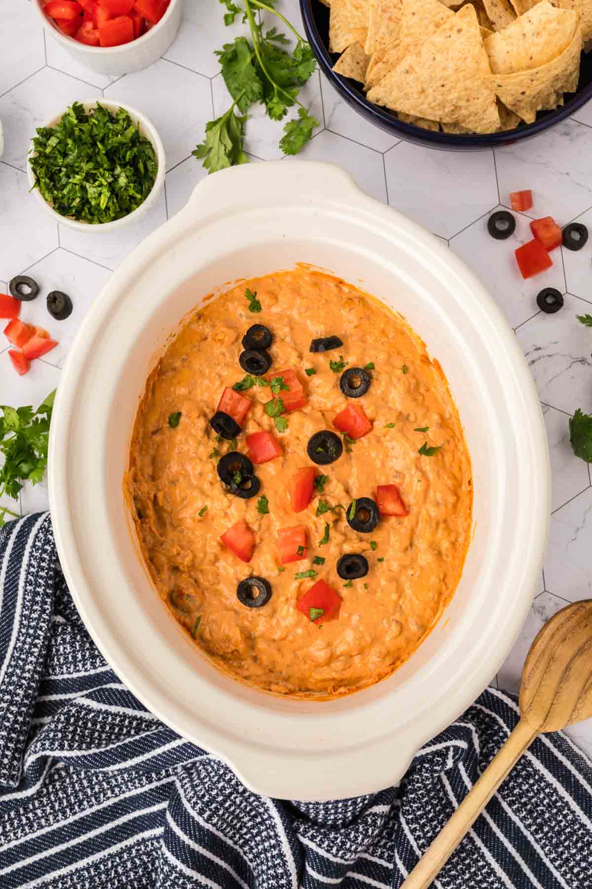 A white ceramic dish filled with creamy orange Crockpot Taco Dip, topped with chopped tomatoes, black olives, and herbs, is surrounded by tortilla chips, cilantro, and diced tomatoes on a white hexagonal surface.