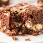 A close-up of a moist chocolate brownie with visible chunks of nuts and a creamy swirl, reminiscent of German chocolate cake cheesecake bars, sitting on a white plate.