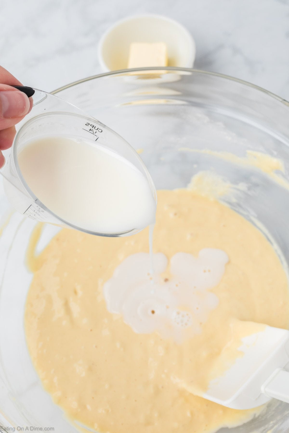 A hand pours milk from a measuring cup into a bowl of yellow batter for Greek Yogurt Pancakes. A white spatula rests inside the bowl, with a small dish of butter in the background on a marble countertop.
