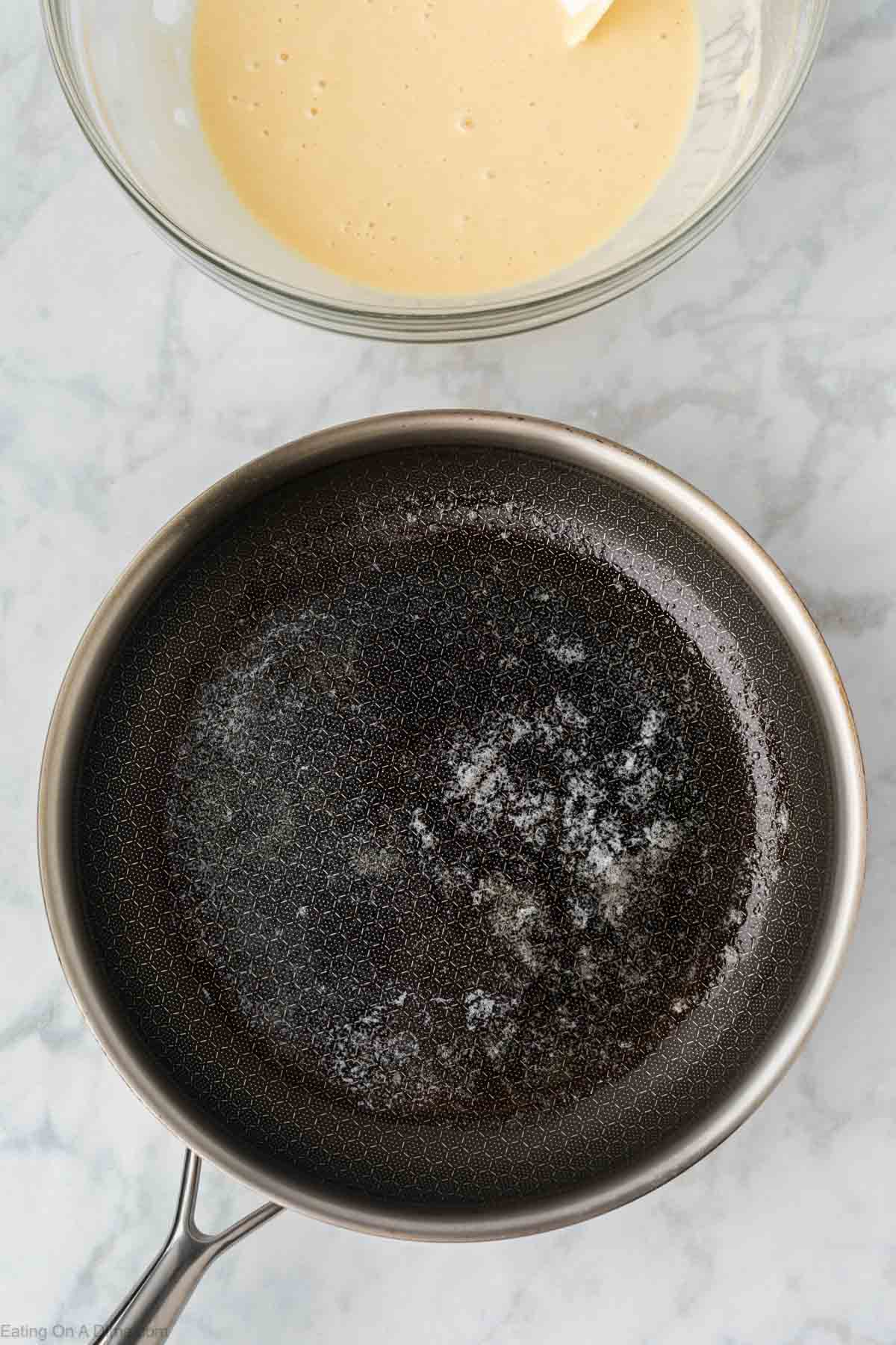 A nonstick frying pan with melted butter sits on a marble countertop, ready for Greek Yogurt Pancakes, with a glass bowl of pancake batter partially visible above it.