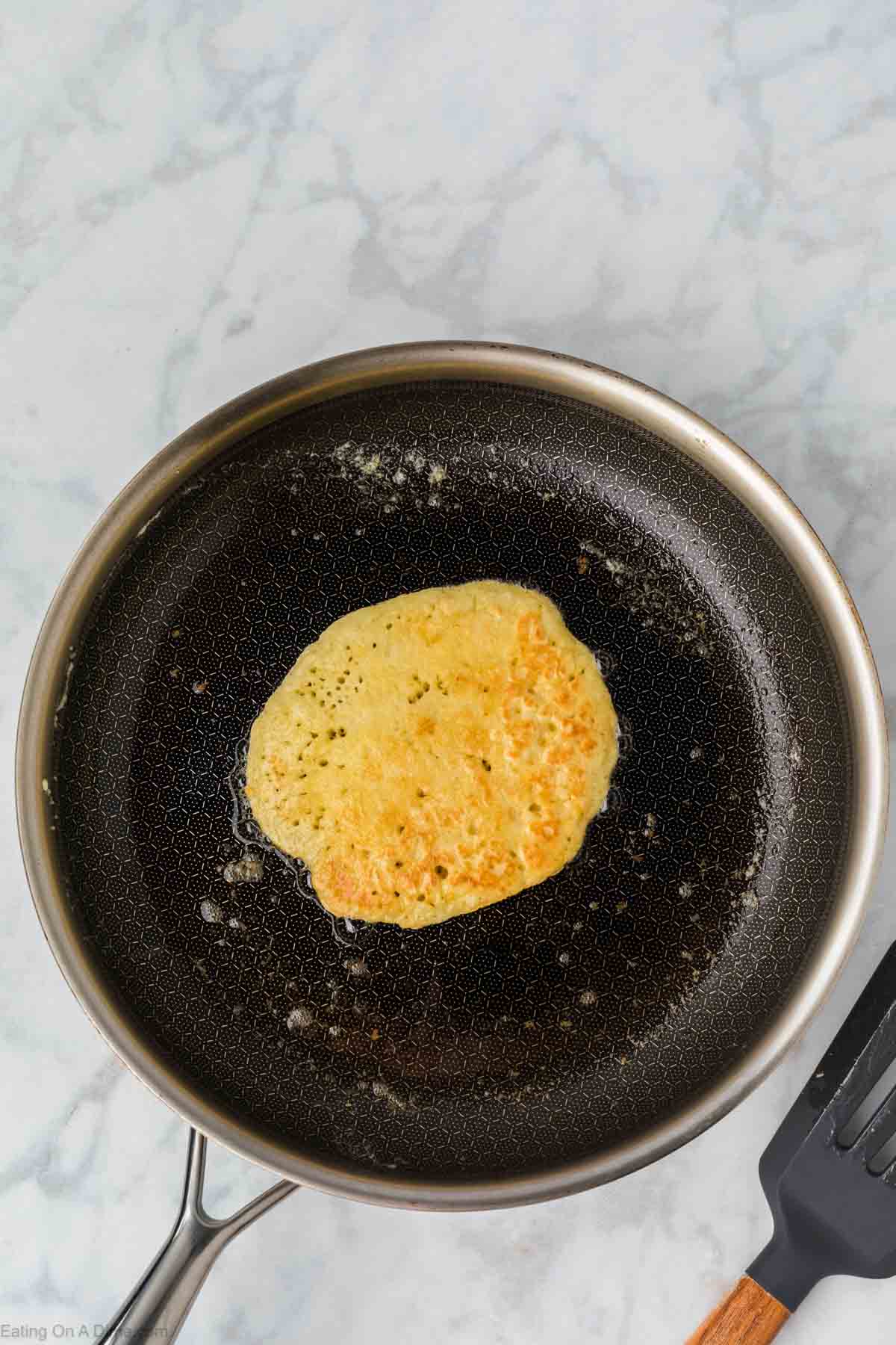A single Greek Yogurt Pancake cooks in a nonstick skillet with melted butter, sitting on a light marble countertop. A black spatula rests nearby.
