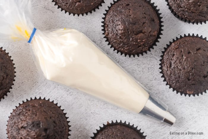 Close up image of a piping bag with marshmallow frosting with several chocolate cupcakes on the side. 