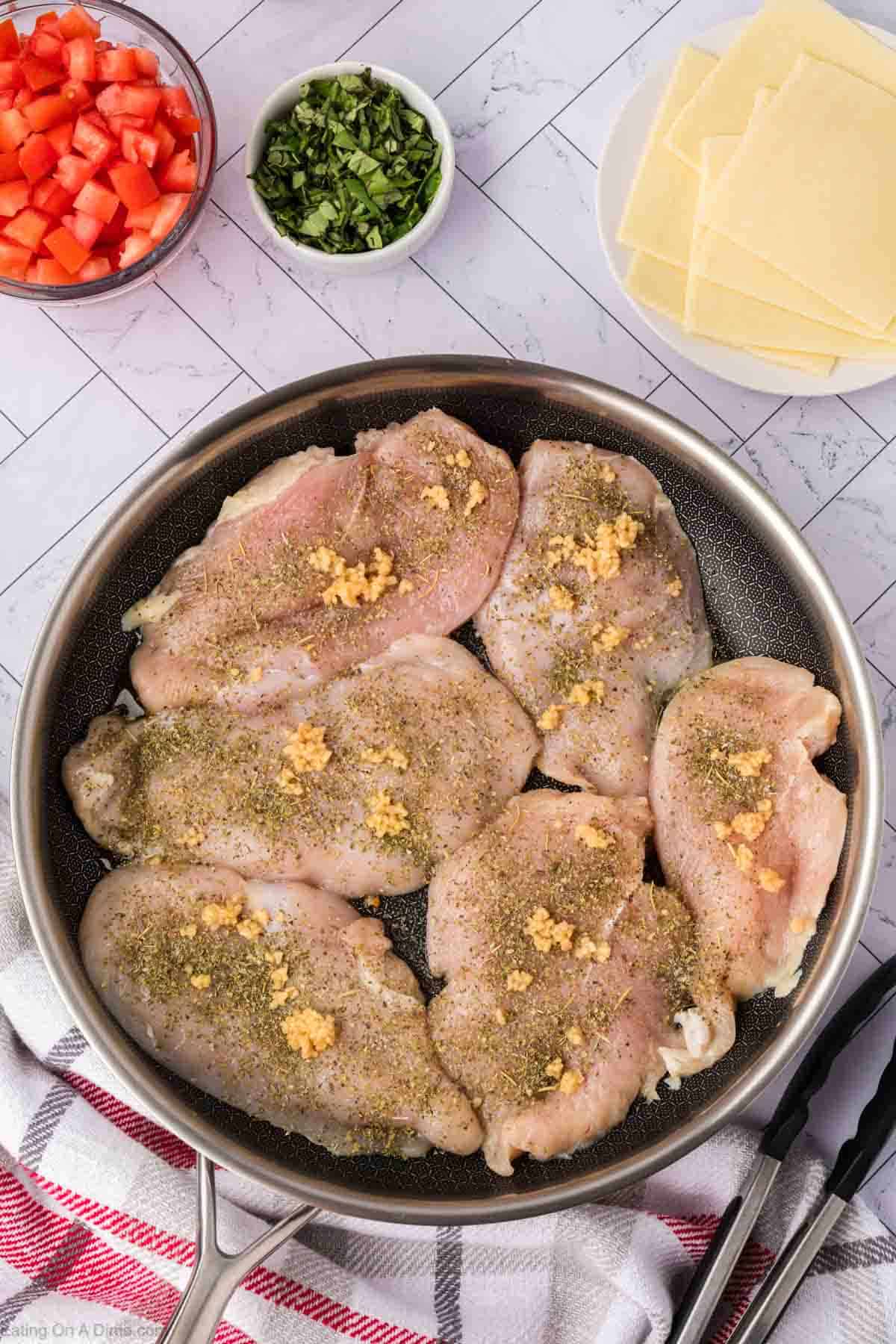 Raw chicken in a skillet topped with seasoning and minced garlic with a small bowl of diced tomatoes, chopped basil and a plate with slice of cheese