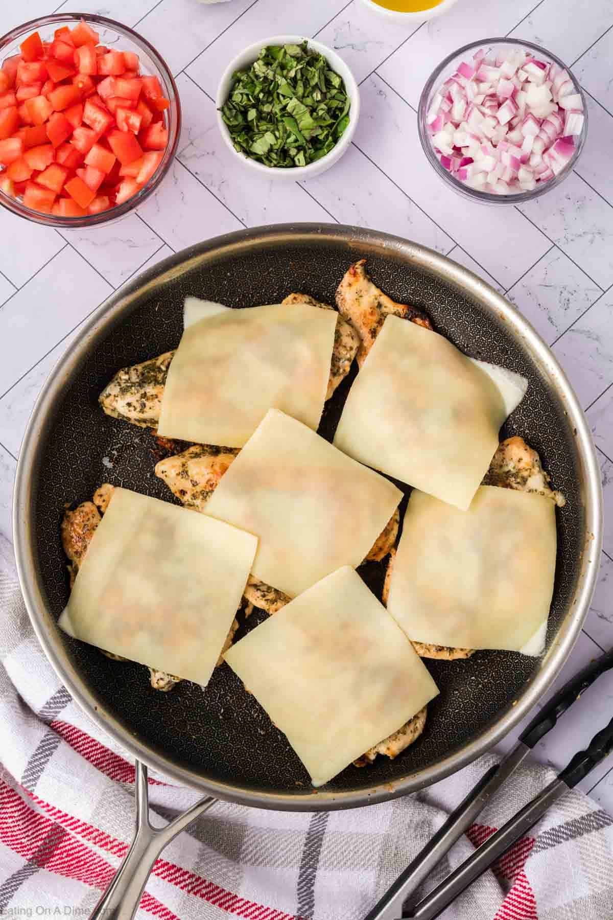 Grilled chicken in a skillet topped with cheese slices with small bowl of diced tomato, chopped basil and red onions