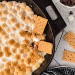 A skillet filled with golden-brown toasted marshmallows creates a tempting s'mores dip, with graham crackers dipped into the mixture. Nearby, broken graham crackers and a bowl of chocolate chips sit on a white wooden surface.